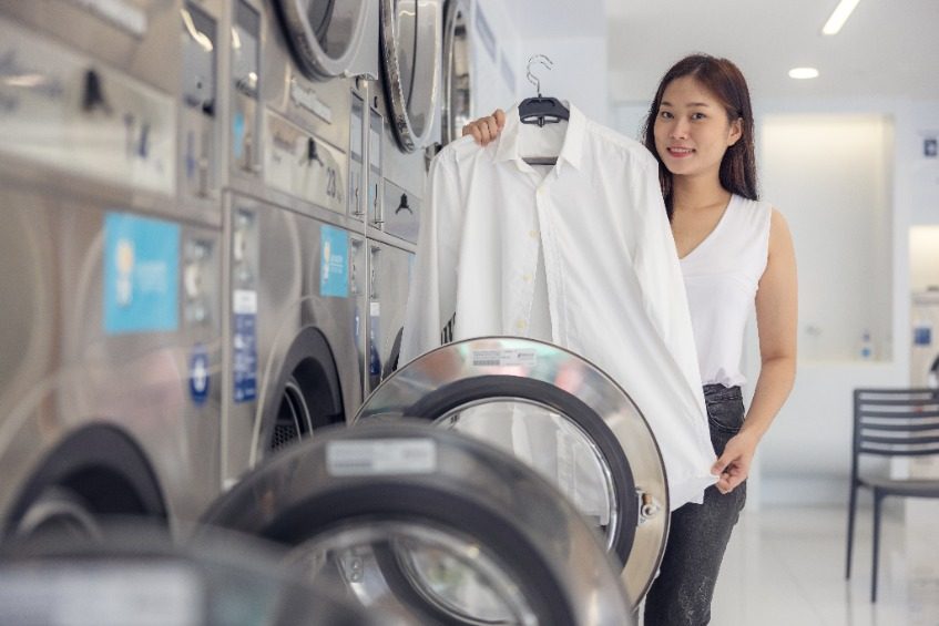 In the self-service laundry with dryer machines in the backdrop, a young woman enjoys clean ironed garments.